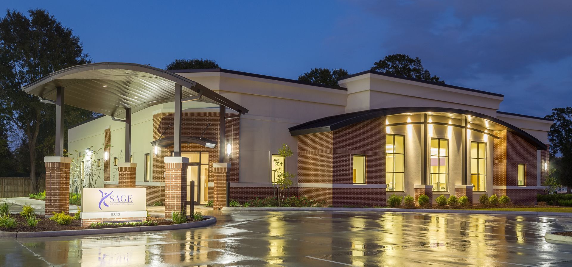 Exterior of a modern building with a curved roof and reflecting wet pavement at dusk.
