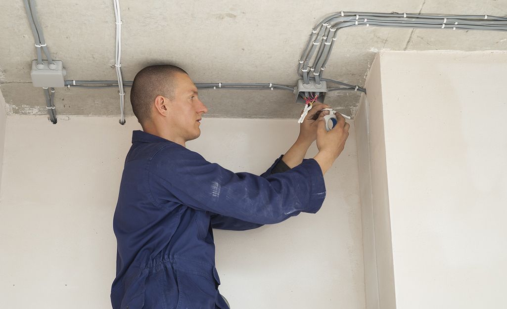 Man in blue jumpsuit installing electrical wiring on a wall and ceiling.