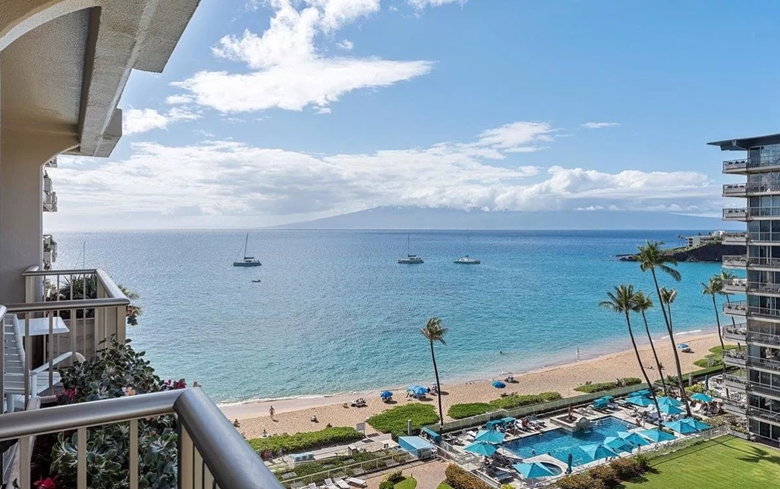 Ocean view from a balcony, beach, pool, blue water, sailboats, palm trees, sunny sky.