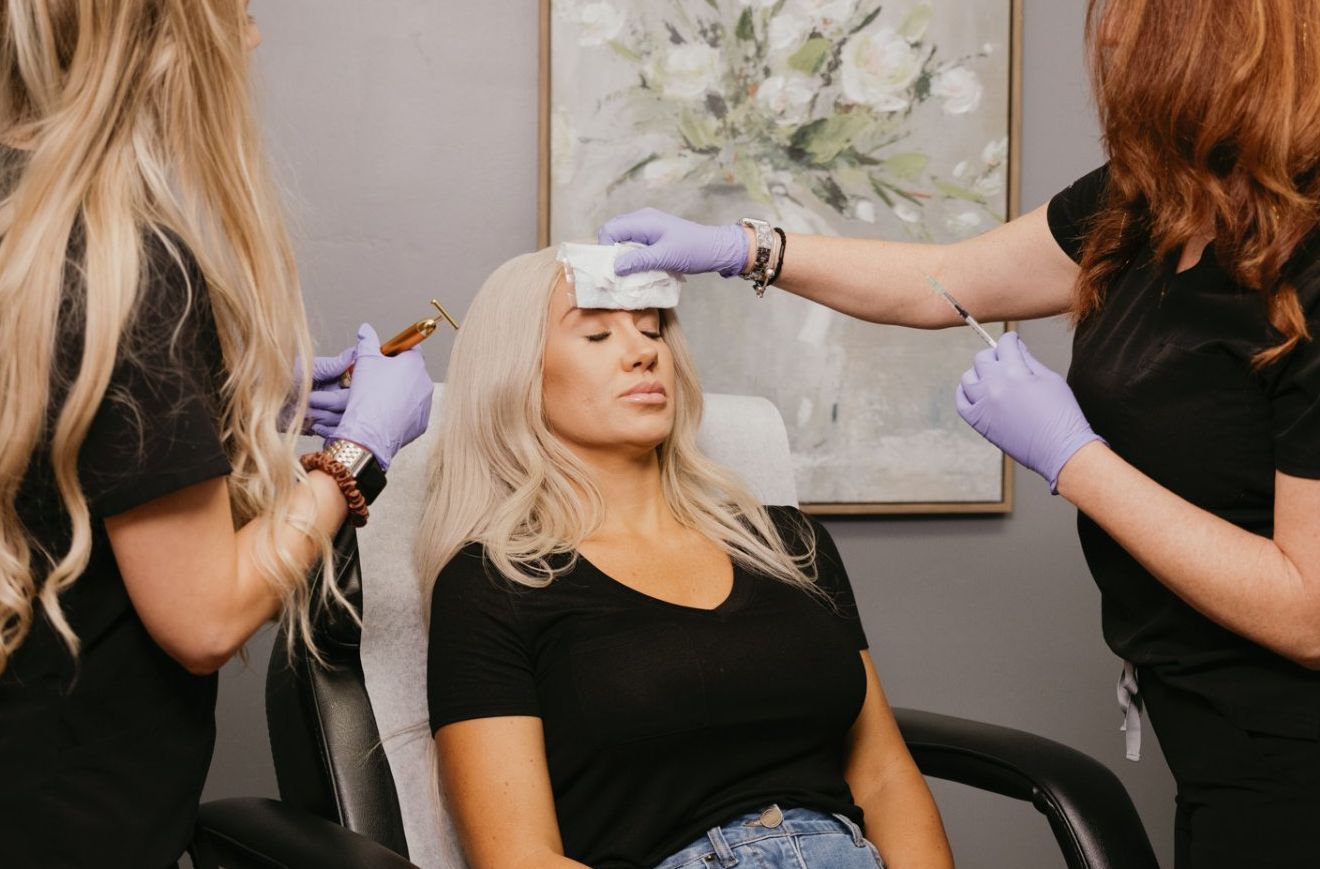 Woman getting a procedure; two people in purple gloves attend to her forehead; salon setting.