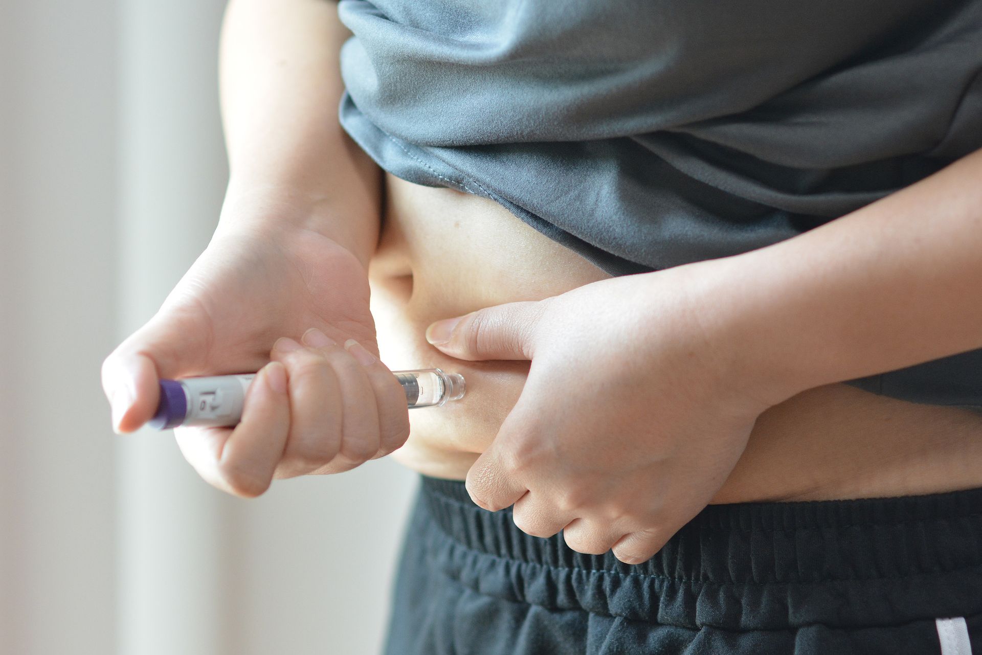 Person injecting insulin into abdomen with a syringe, wearing a gray shirt and black pants.