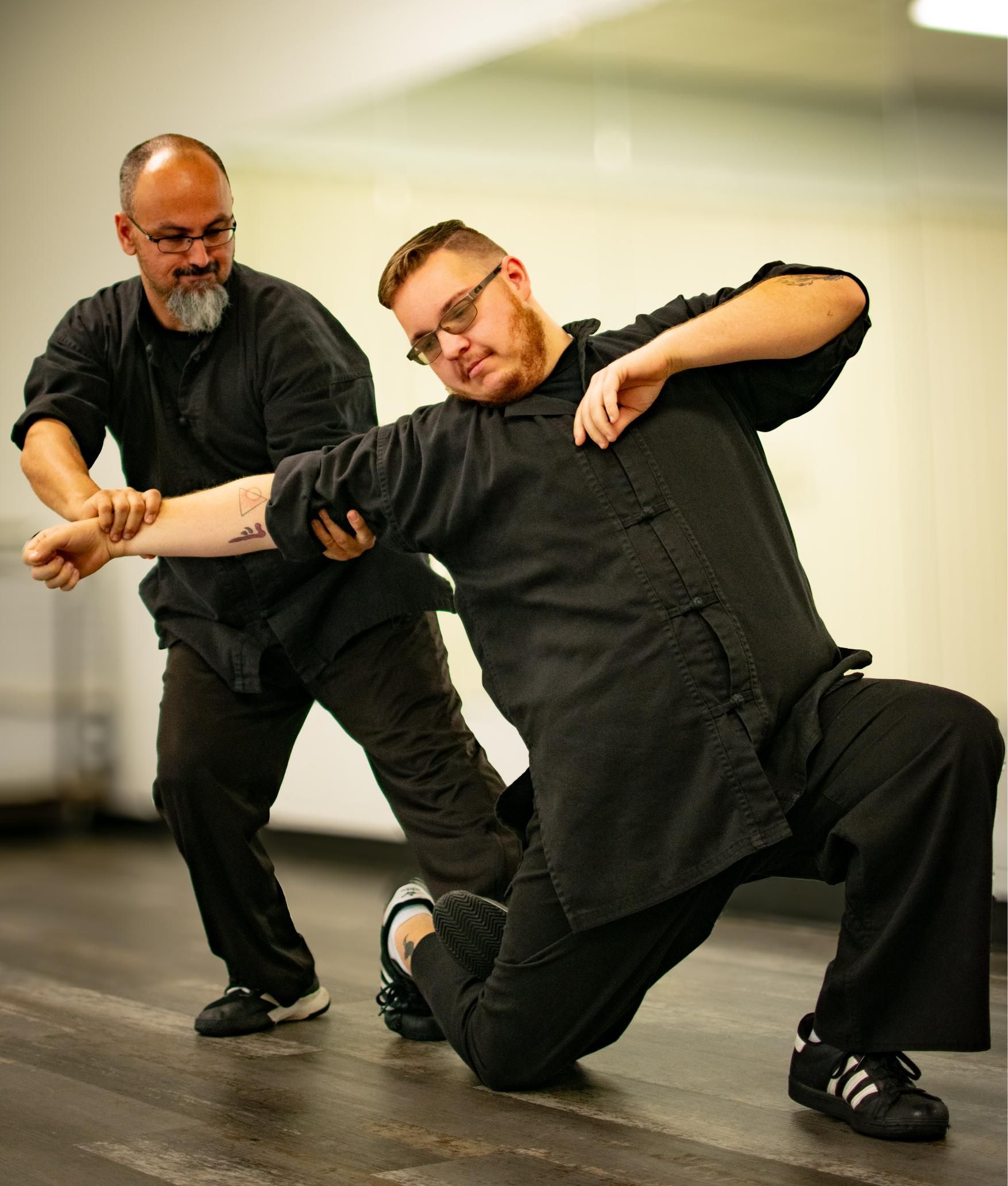 Two men in black martial arts uniforms practicing self-defense in a studio. One is grabbing the other’s arm.