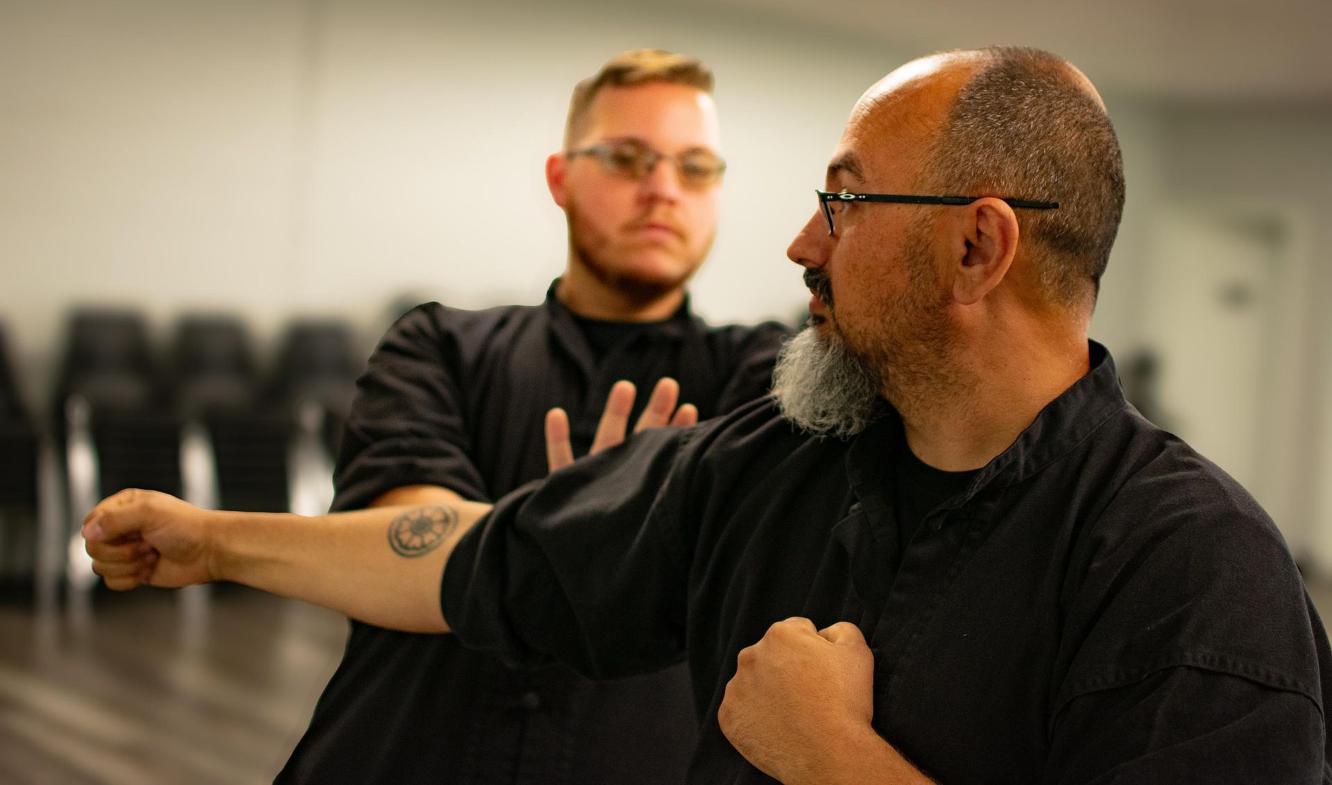 Two men practicing martial arts. Older man in black shirt punches, younger man watches with hands up.