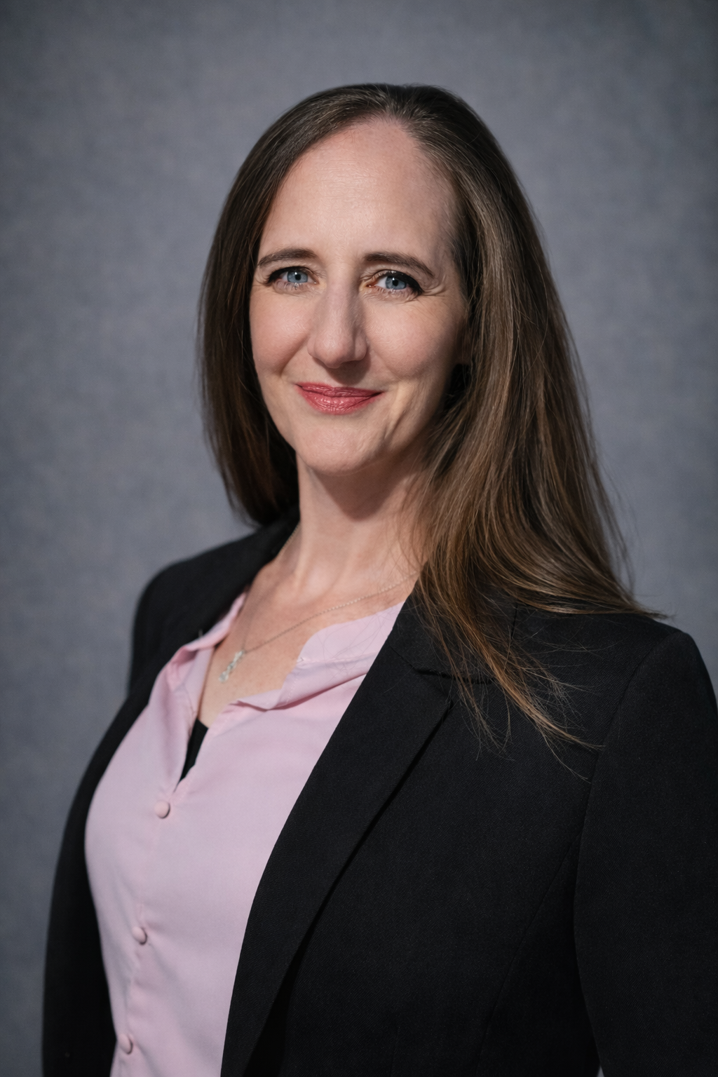 Woman with long brown hair, wearing a pink shirt and black blazer, smiling in front of a gray backdrop.