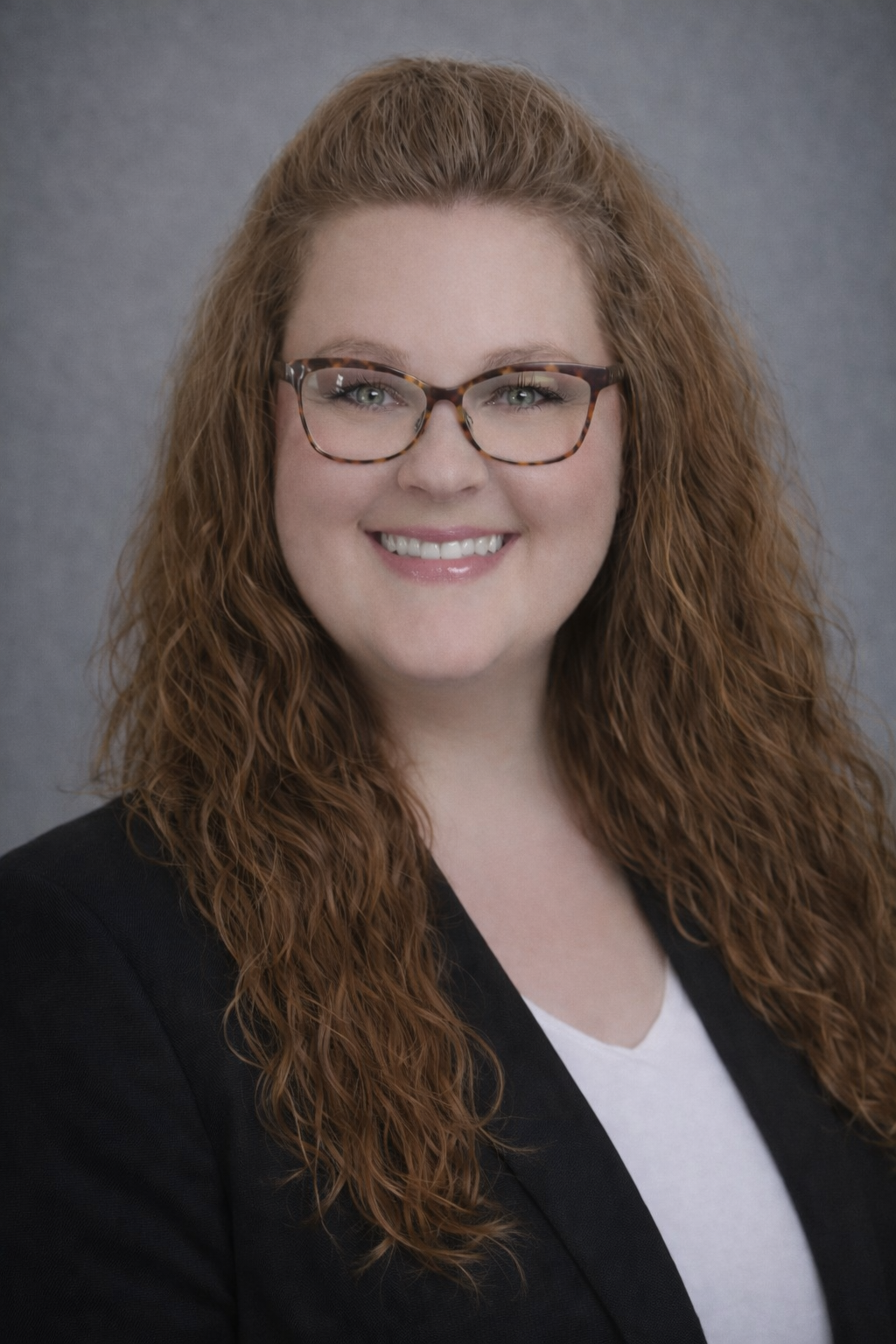 Woman with curly red hair, glasses, and a blazer smiles at the camera.