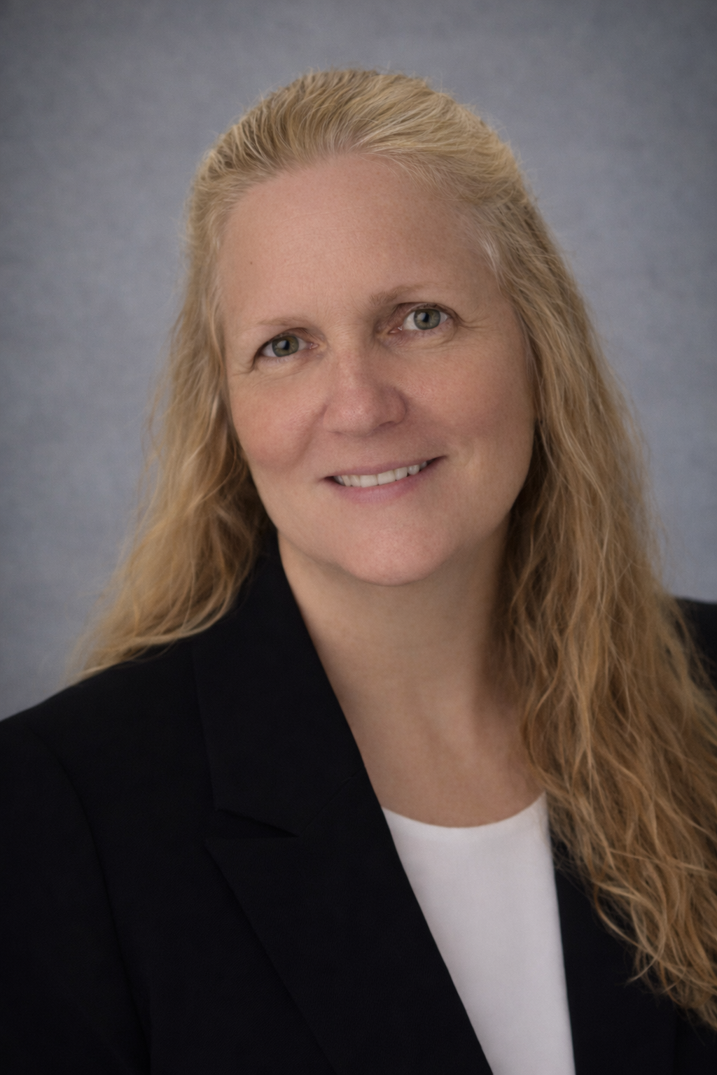 Woman with blonde hair wearing a black blazer smiles at the camera against a blue background.