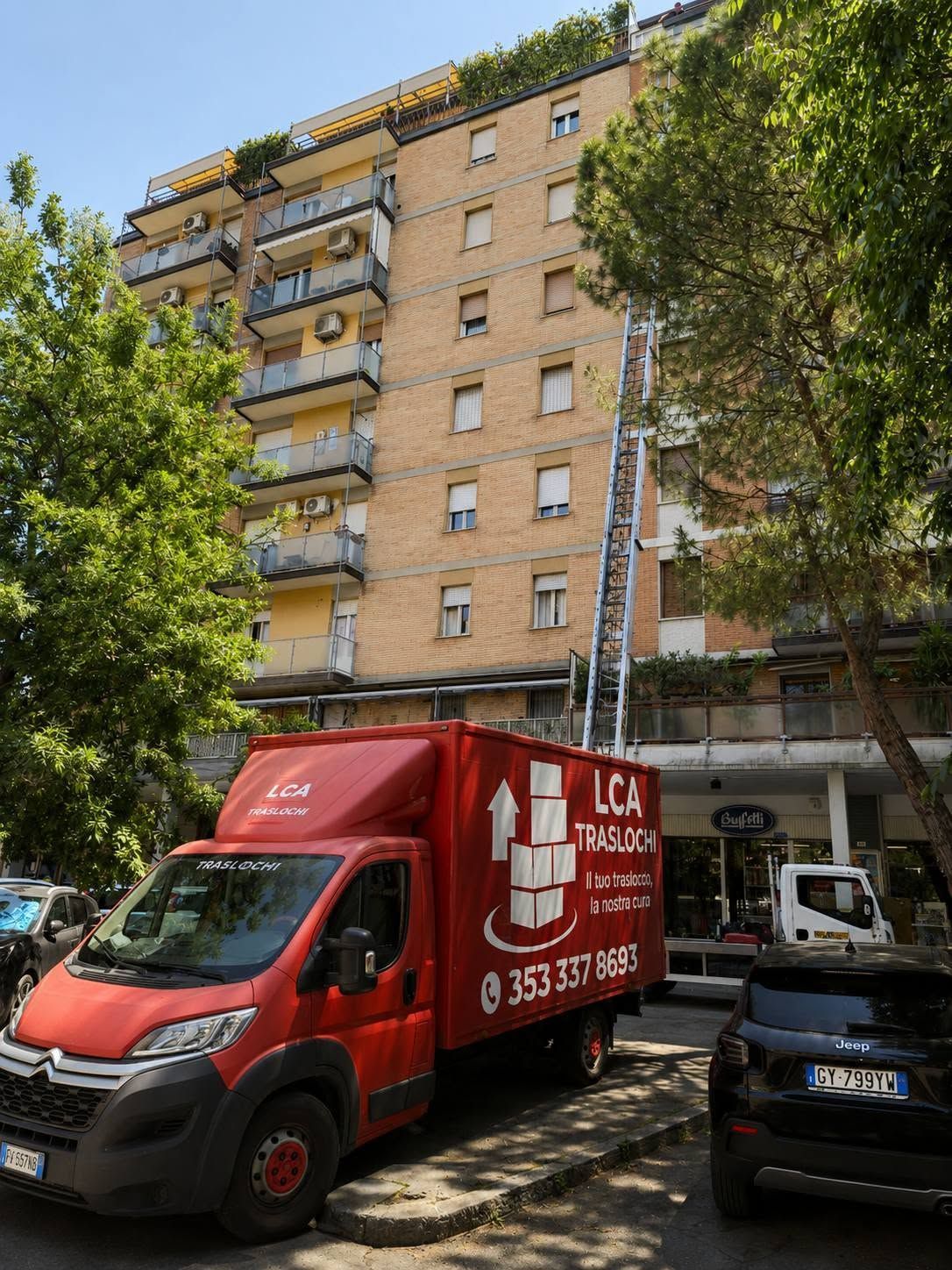 Un camioncino rosso per le consegne è parcheggiato in una strada cittadina alberata, accanto a un condominio.