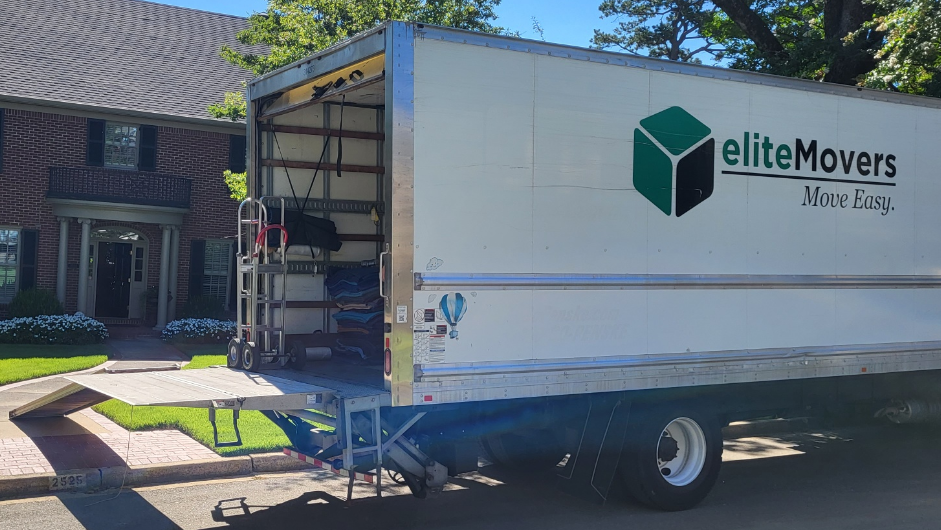 An Elite Movers moving truck parked on a residential street with its back ramp lowered and a dolly inside.