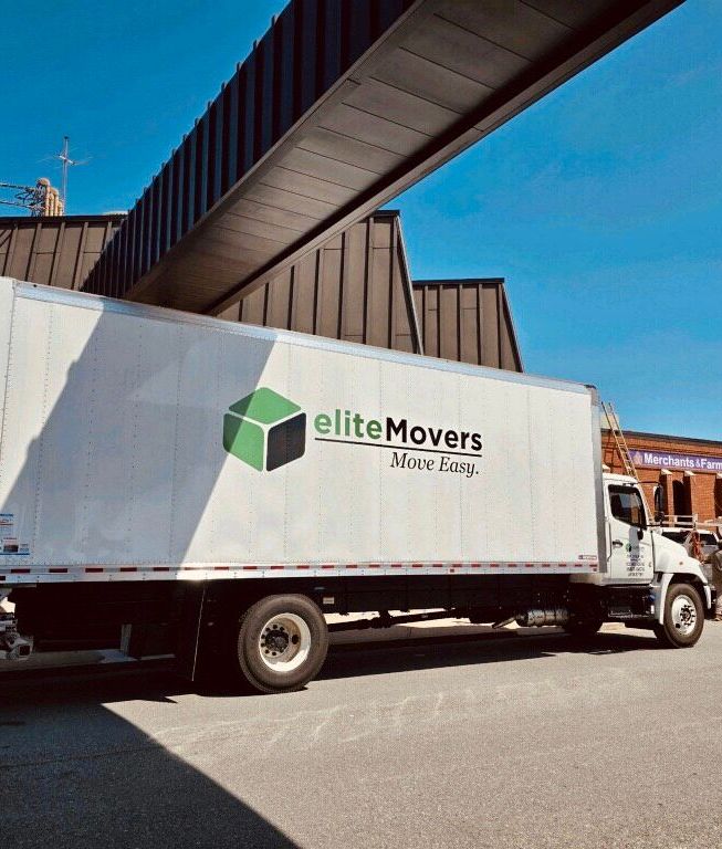White Elite Movers truck parked under an elevated roadway on a sunny street.