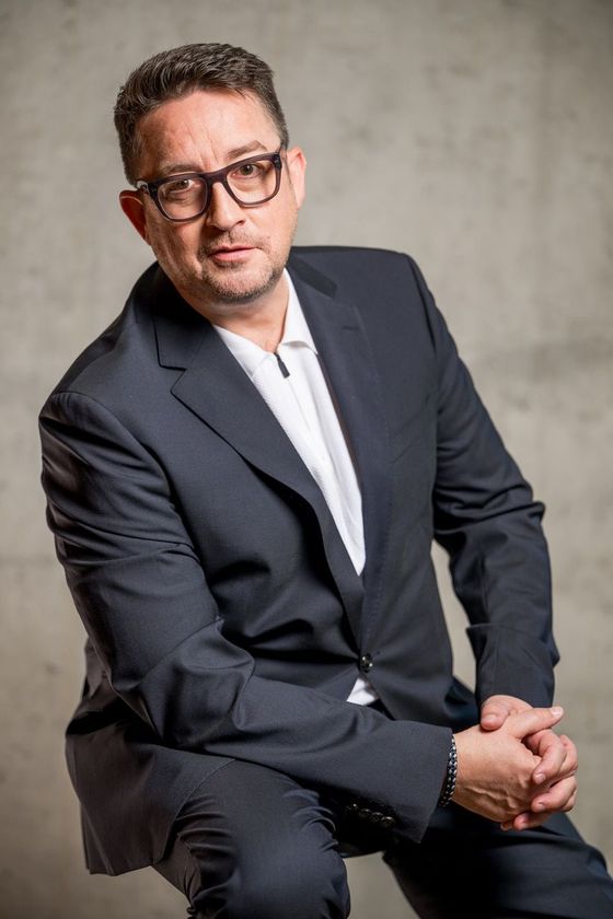 Mann in dunklem Anzug und Brille sitzt vor einem grauen Studiohintergrund, die Hände verschränkt.