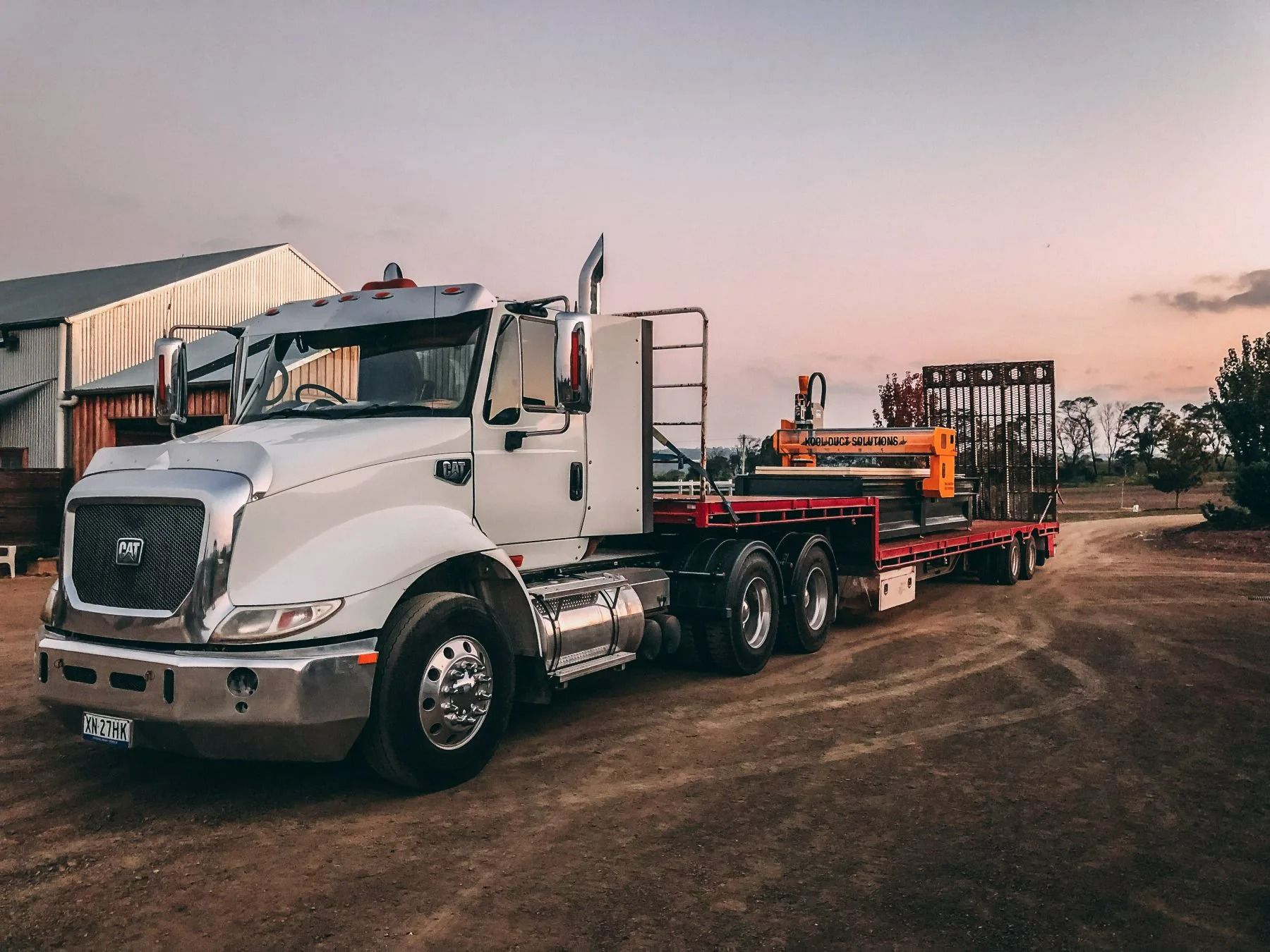 White semi-truck with flatbed trailer on a dirt road near a barn at dusk — Highlands Water Cartage in Glenquarry, NSW