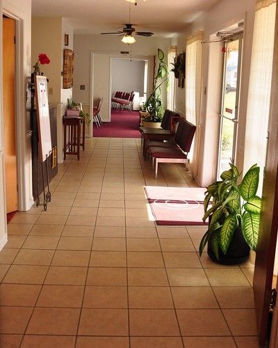 Long hallway with tiled floor, chairs, potted plants, and a red rug leading to a bright room.