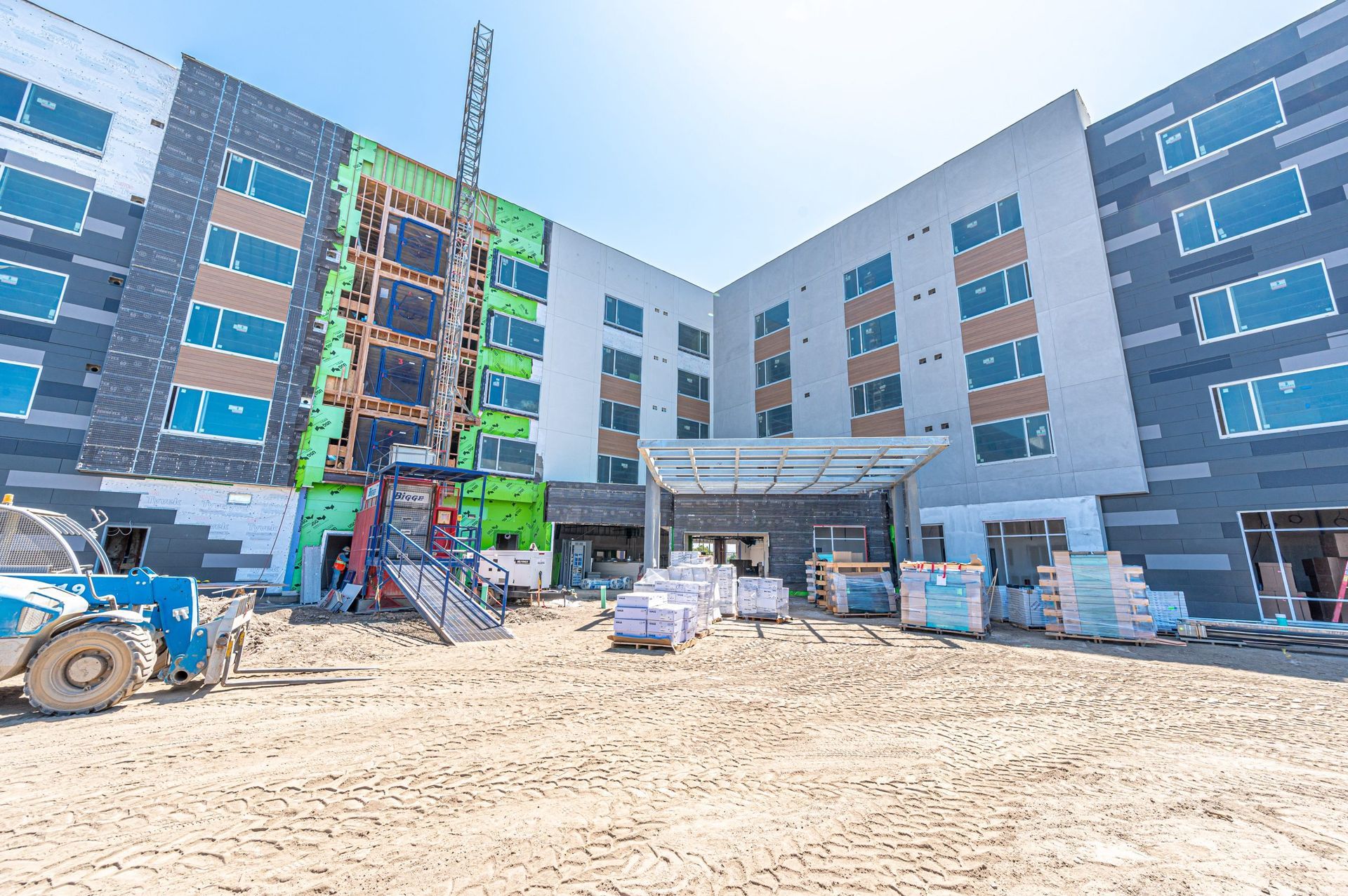 A multi-story hotel building under construction, featuring gray siding, exposed green panels, and a dirt-covered ground.