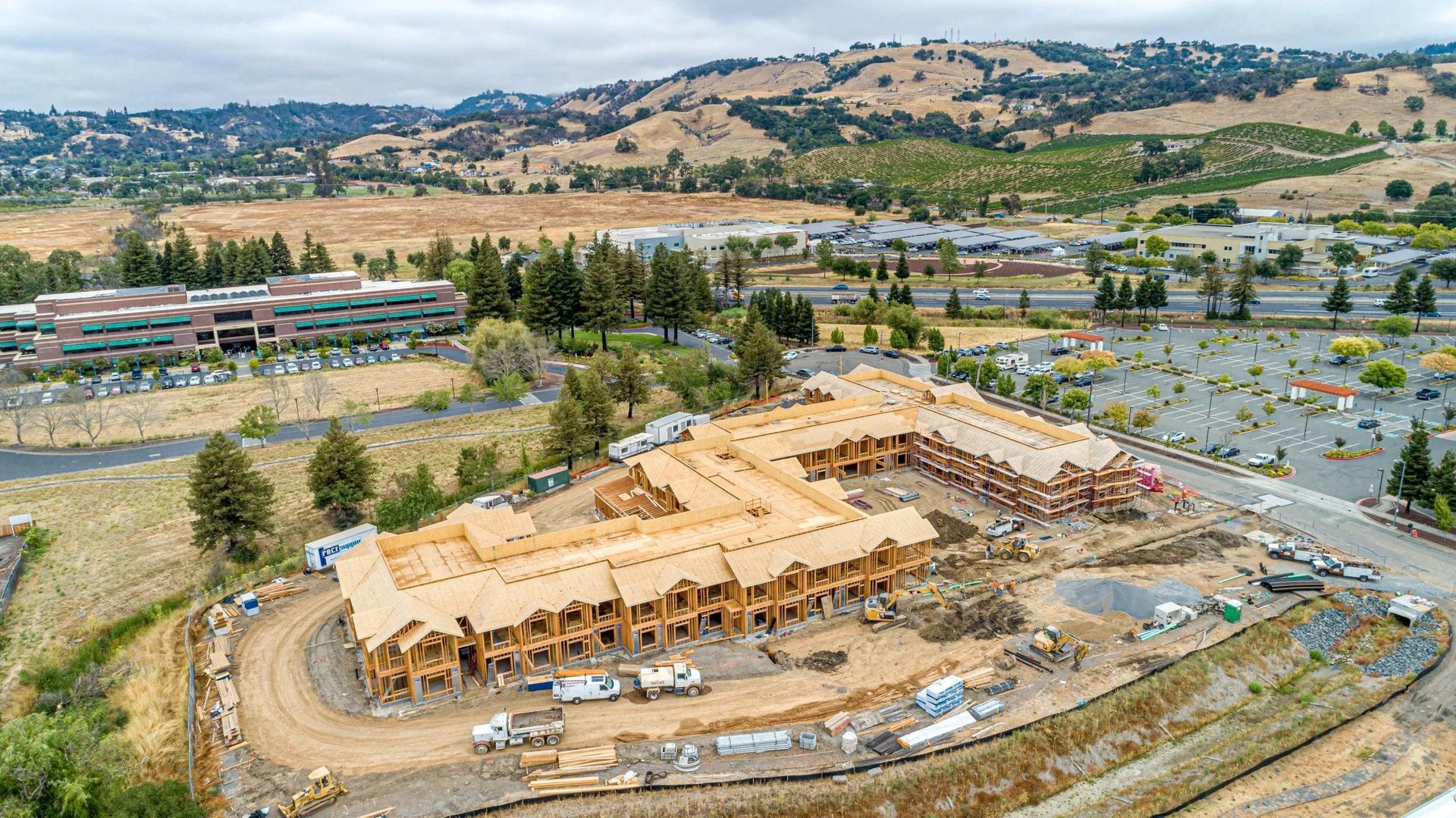 Aerial view of a wooden townhouse complex under construction on a dirt lot, surrounded by trees, parking, and hills.