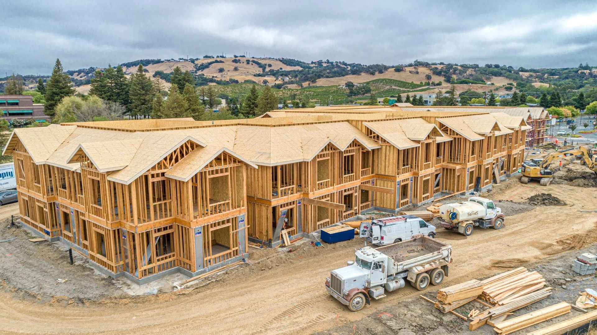 A wooden apartment complex under construction with a dump truck parked on the dirt site in a hilly, rural landscape.