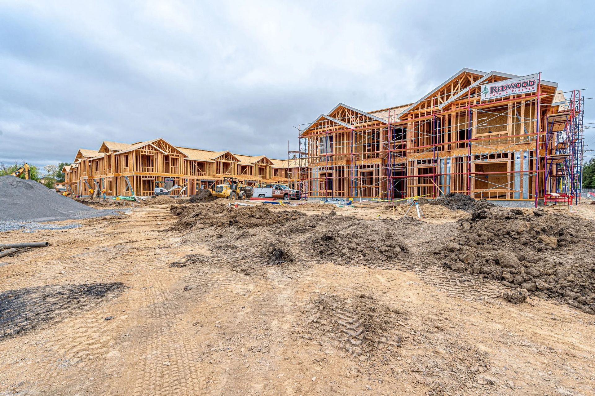 Wood-framed houses under construction on a muddy residential lot with overcast sky