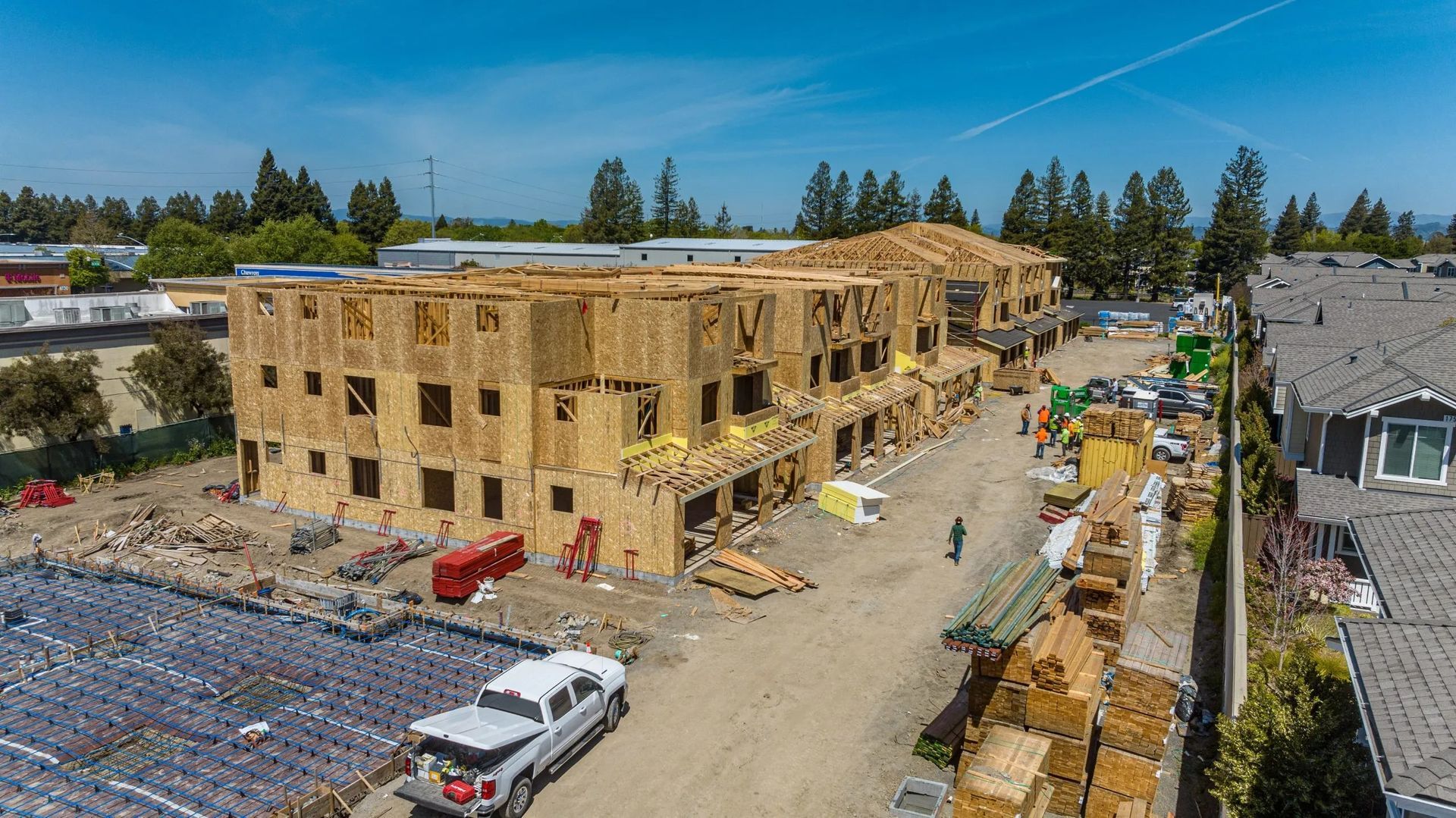 Wood-framed apartment building under construction, with workers, vehicles, and materials on a sunny site