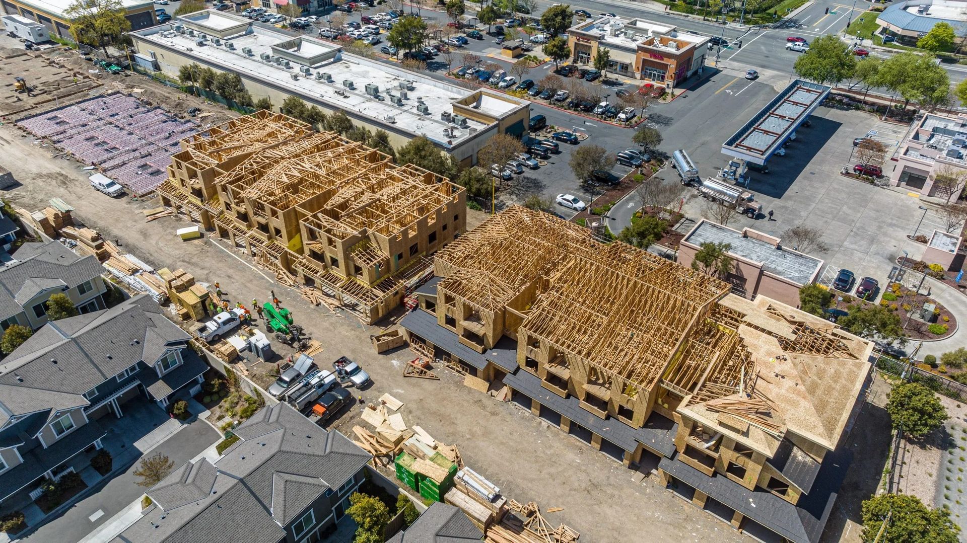 An aerial view shows two multi-story wooden building frames under construction next to a parking lot and existing homes.