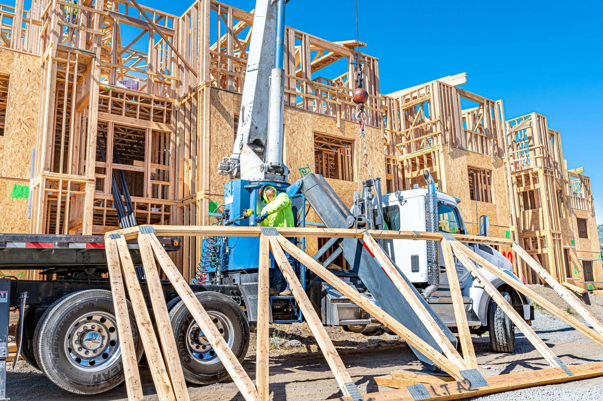 A mobile crane on a truck bed delivers wooden roof trusses to a multi-story wood-frame building under construction.