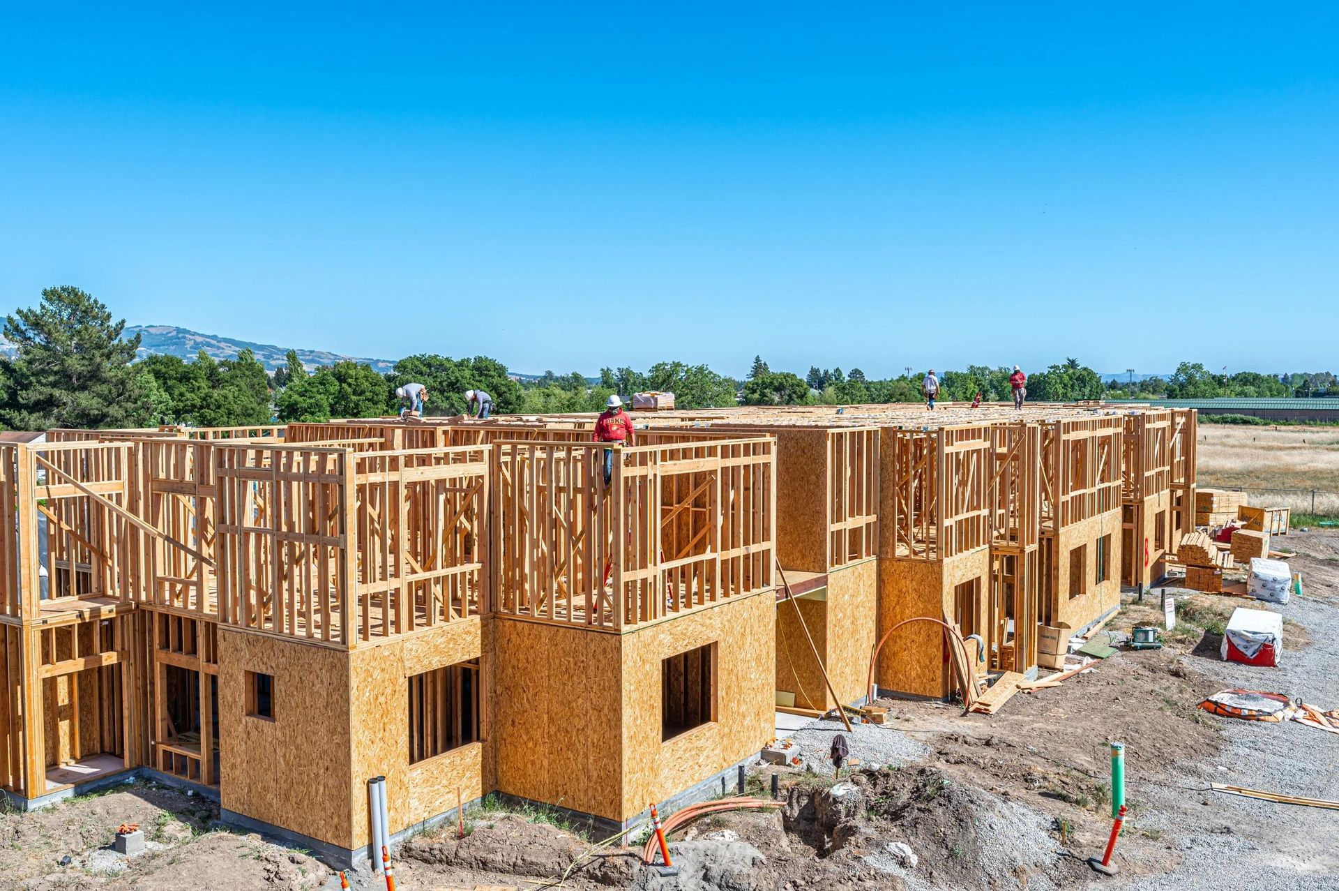 Partially built wooden townhouses under construction on a gravel site under a clear blue sky