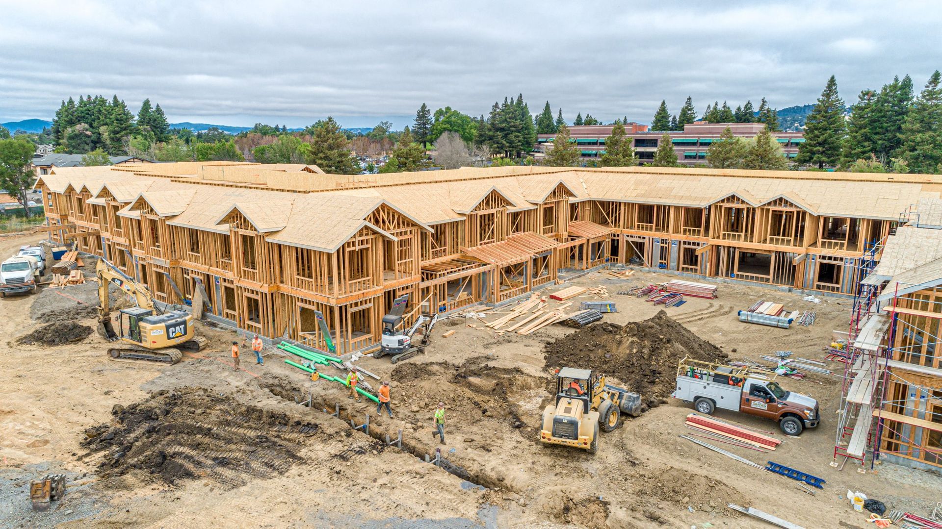 An aerial view of a construction site featuring a large, multi-unit wooden building frame under a cloudy sky.