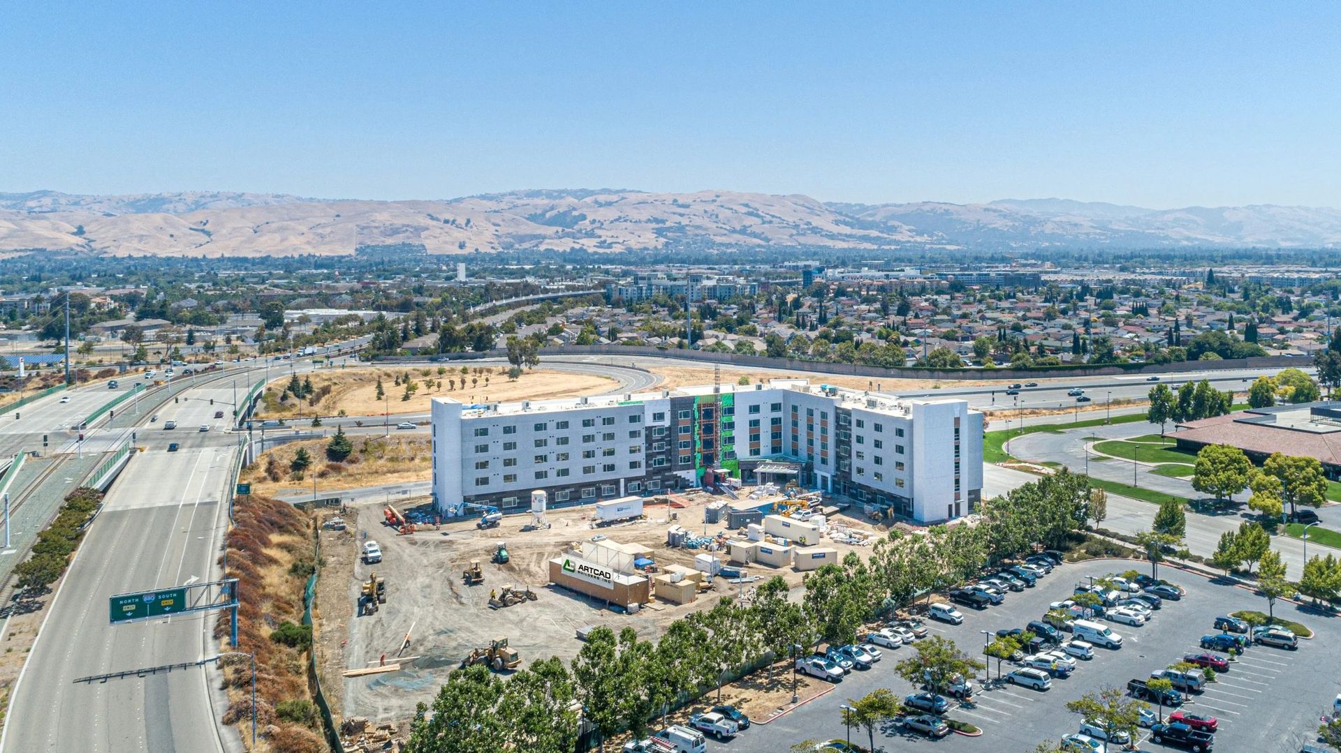 Aerial view of a multistory building under construction next to a highway and a parking lot with mountains in the distance.