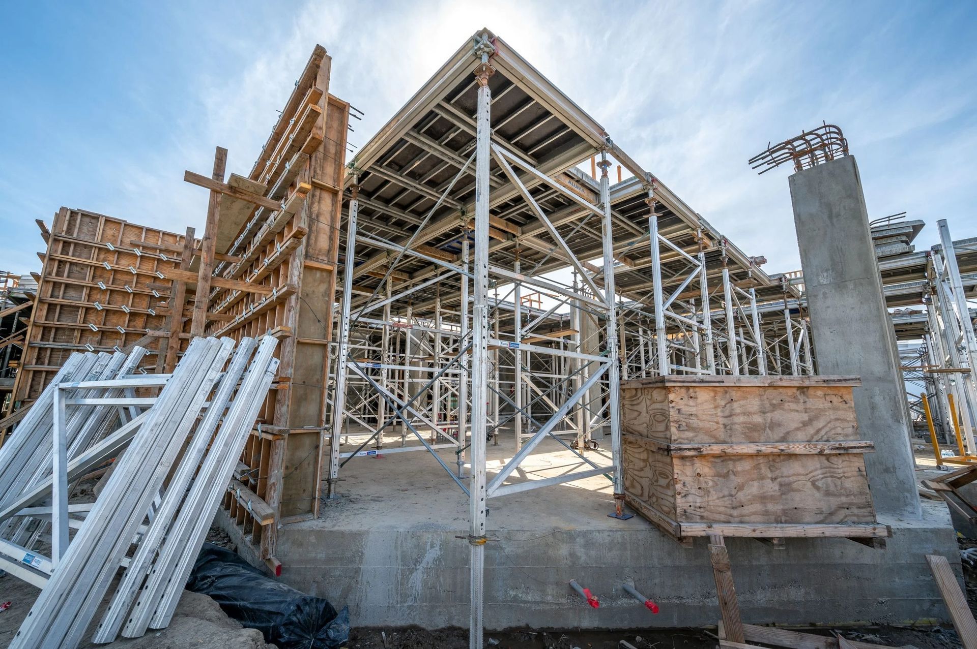 Construction site showing scaffolding and wooden formwork for concrete floor slabs beneath a bright, sunny blue sky.