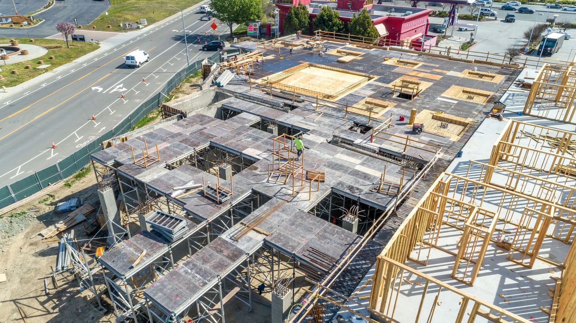 An aerial view of a construction site with concrete flooring, structural framing, and nearby roads and buildings.