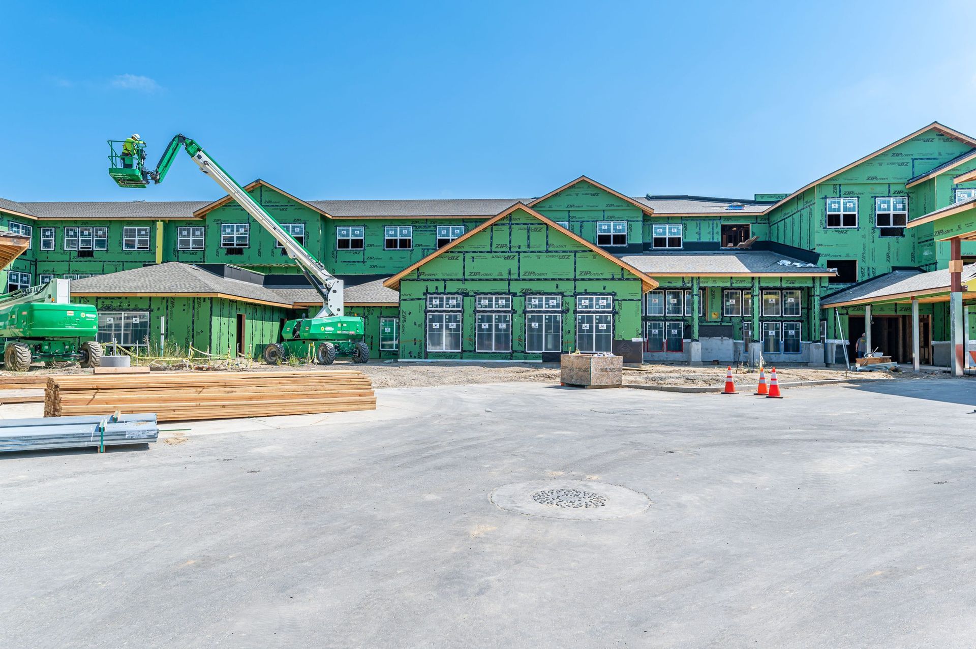 Construction site with a green-sheathed building, a boom lift, and stacks of lumber on a sunny day.