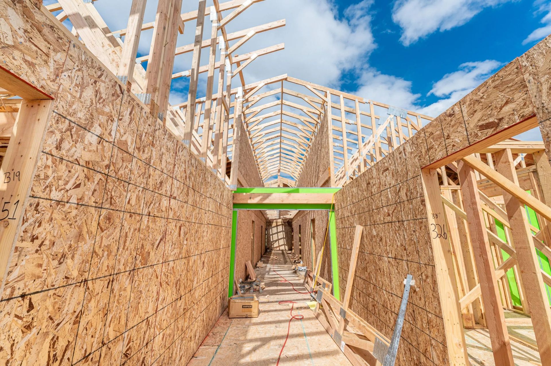 Low-angle view of a wood-framed house under construction, showing plywood walls, roof trusses, and a bright green beam.