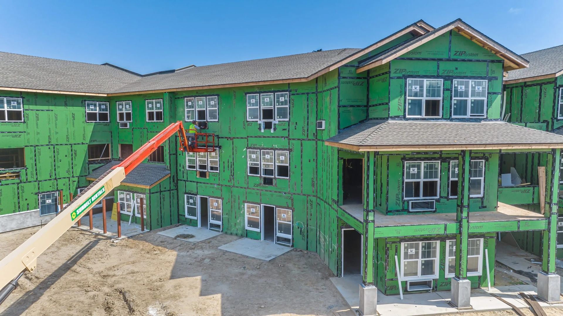 A multi-story apartment building under construction, covered in green sheathing, with an orange construction lift outside.