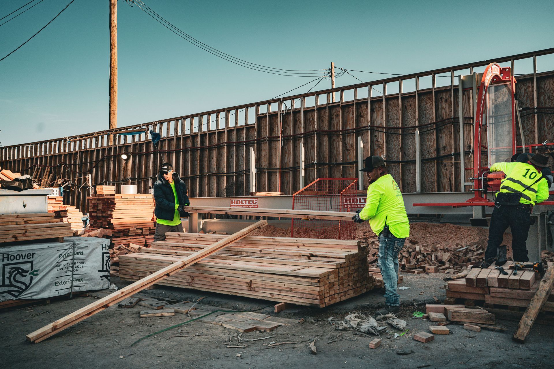 Construction workers in neon jackets handle lumber at an outdoor jobsite beside a concrete wall.