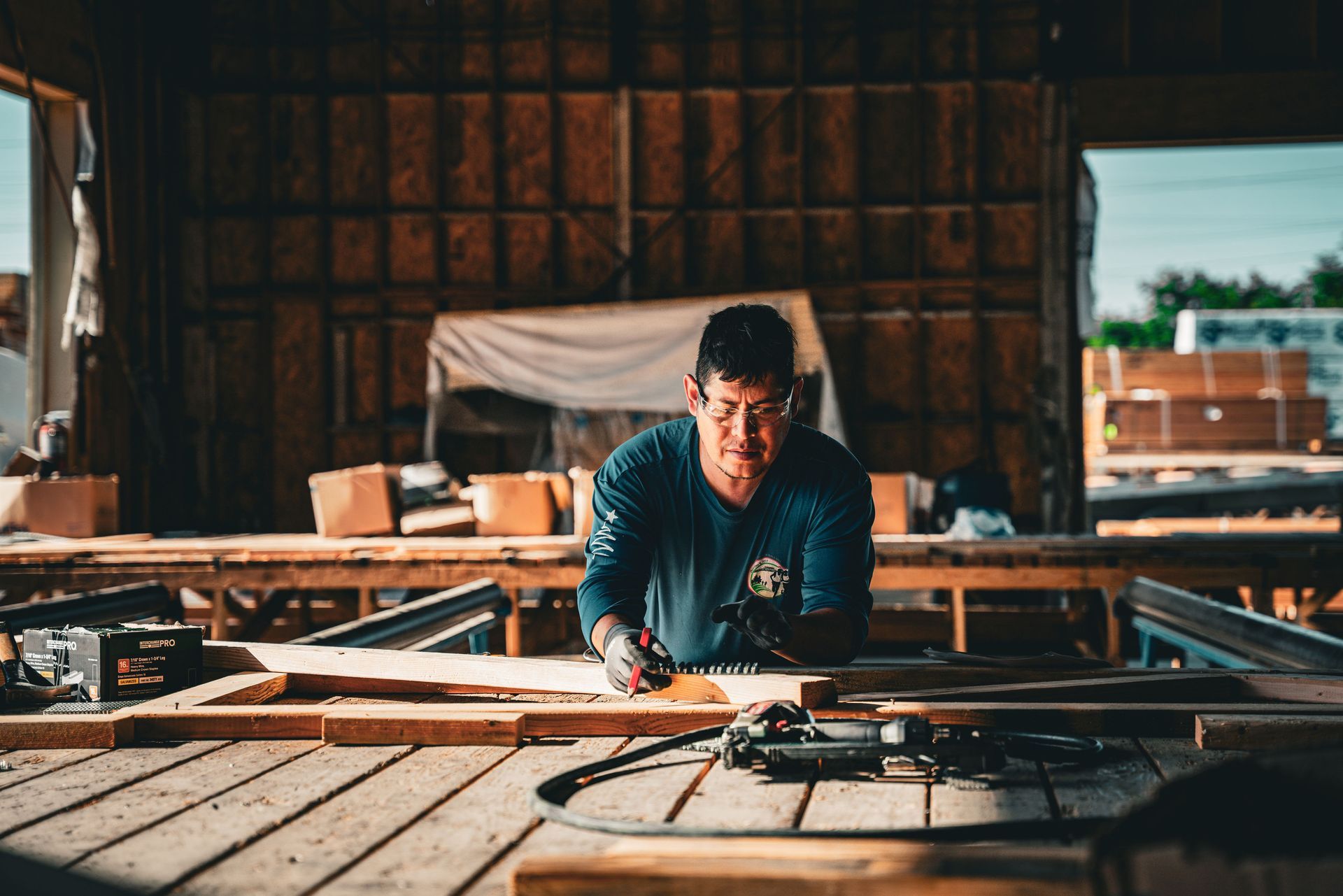 Person working on a wooden frame in a workshop, leaning over a table with tools and materials.