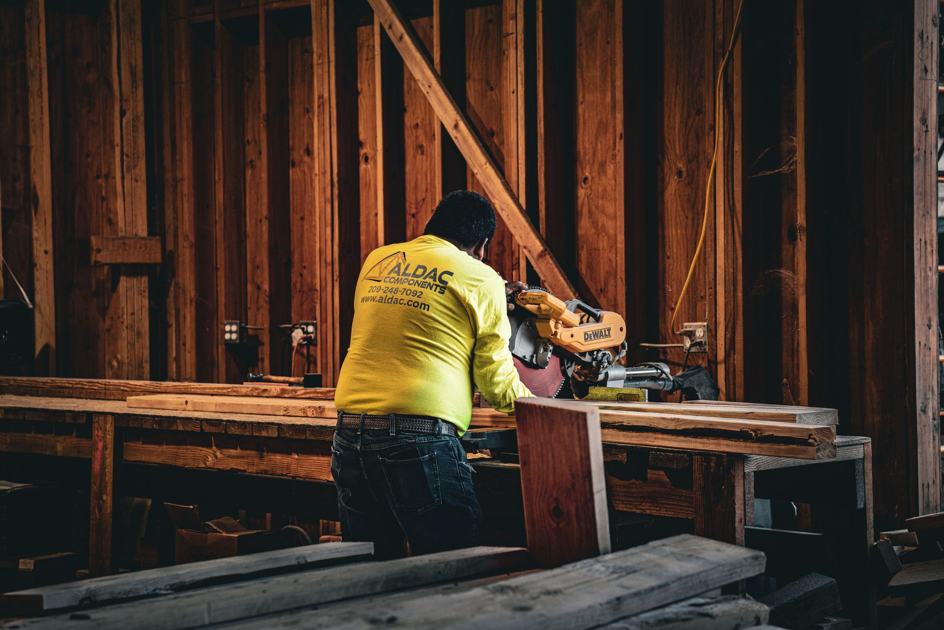 Person in a yellow shirt uses a power tool on a wooden workbench inside a dim carpentry shop