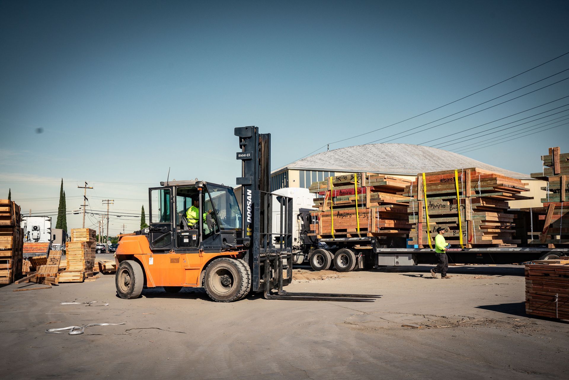 Orange forklift lifting stacked pallets in an industrial yard with workers and lumber in the background