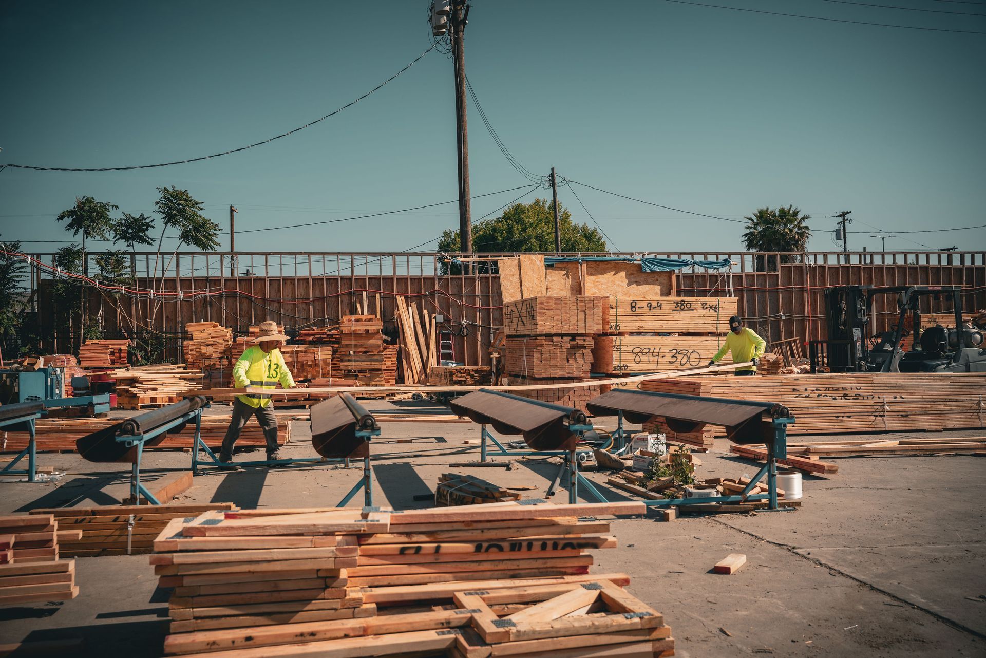 Wooden pallets and lumber stacked in a yard, with workers in safety vests near construction materials.