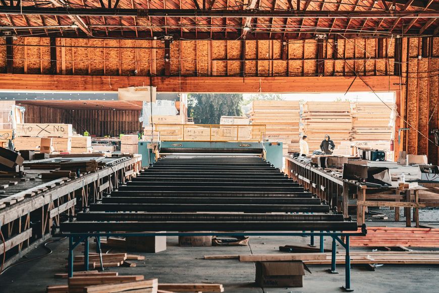 Stacked black metal beams in an open-sided workshop with a thatched roof and wooden tables.