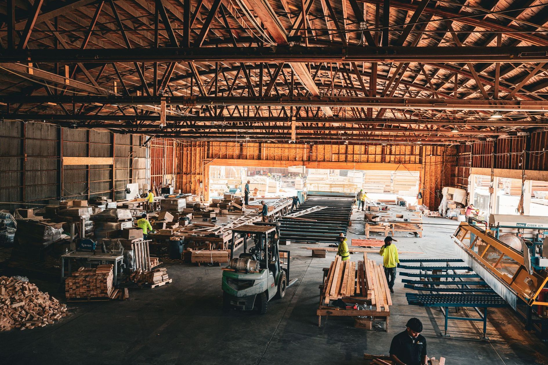 Busy lumber mill interior with workers, stacked wood, and large industrial machinery under a high wooden roof