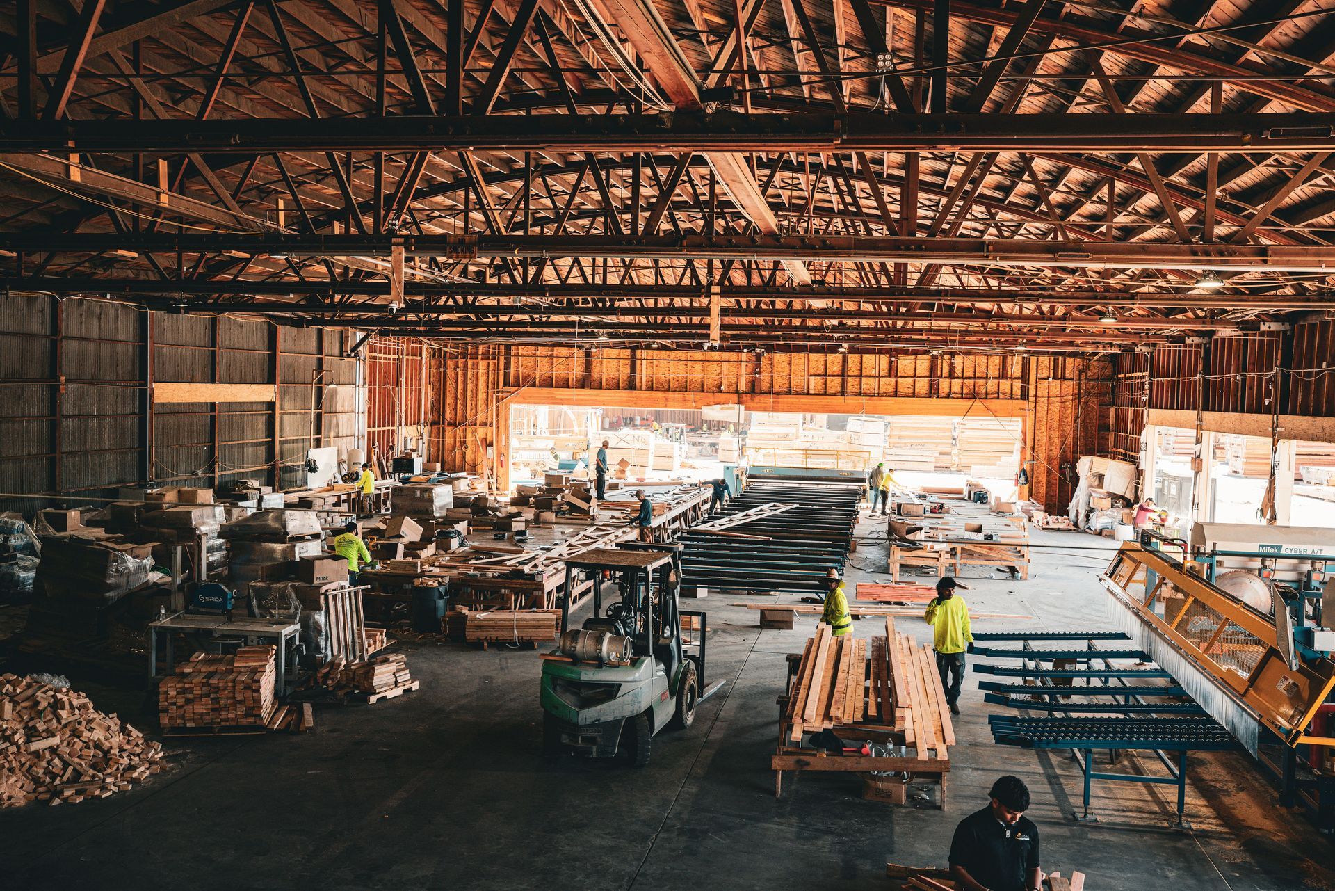 Busy lumber mill interior with workers, stacked wood, and large industrial machinery under a high wooden roof