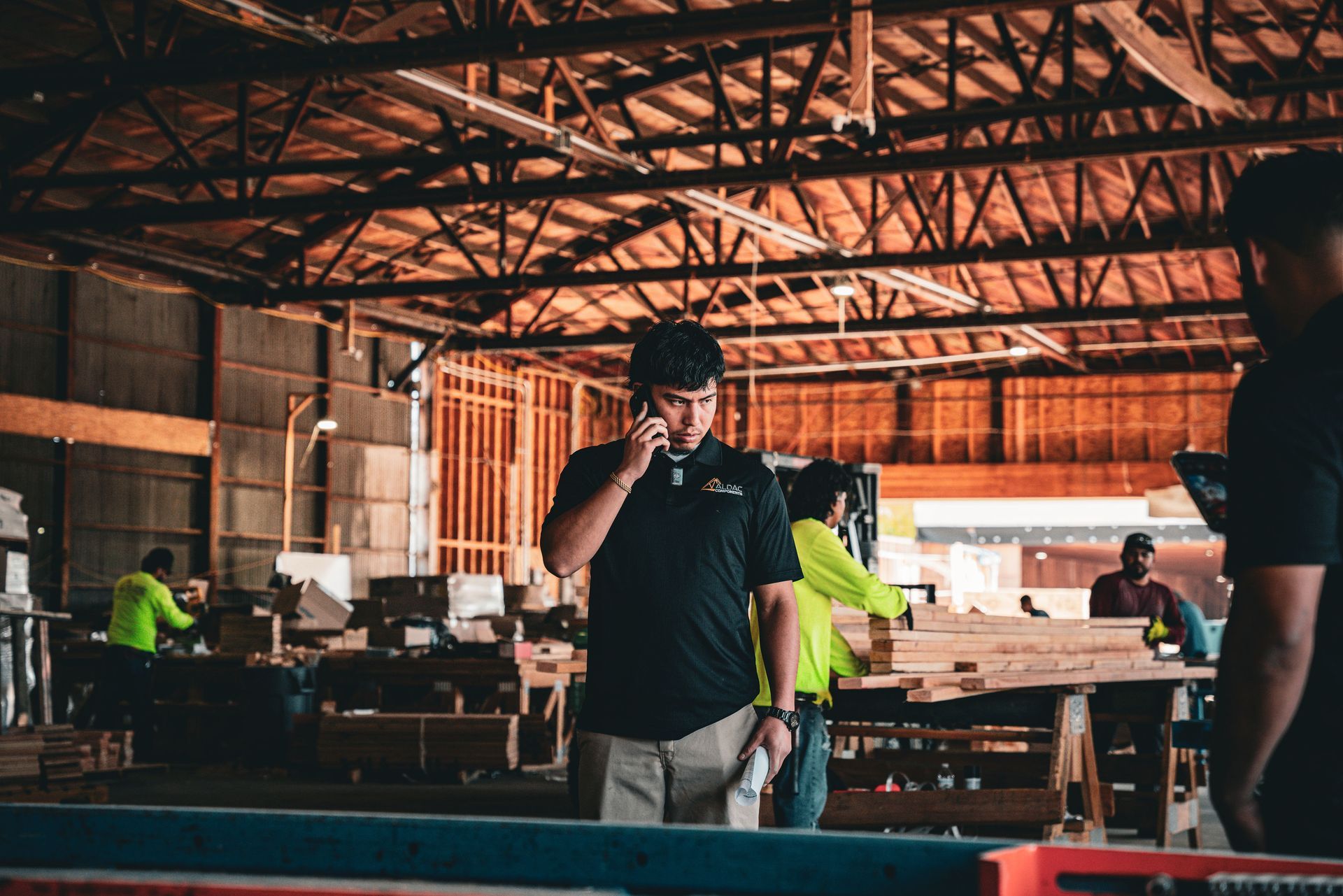 Workers in a warehouse assembling wooden tables under exposed beams, with a man in a black shirt in the foreground