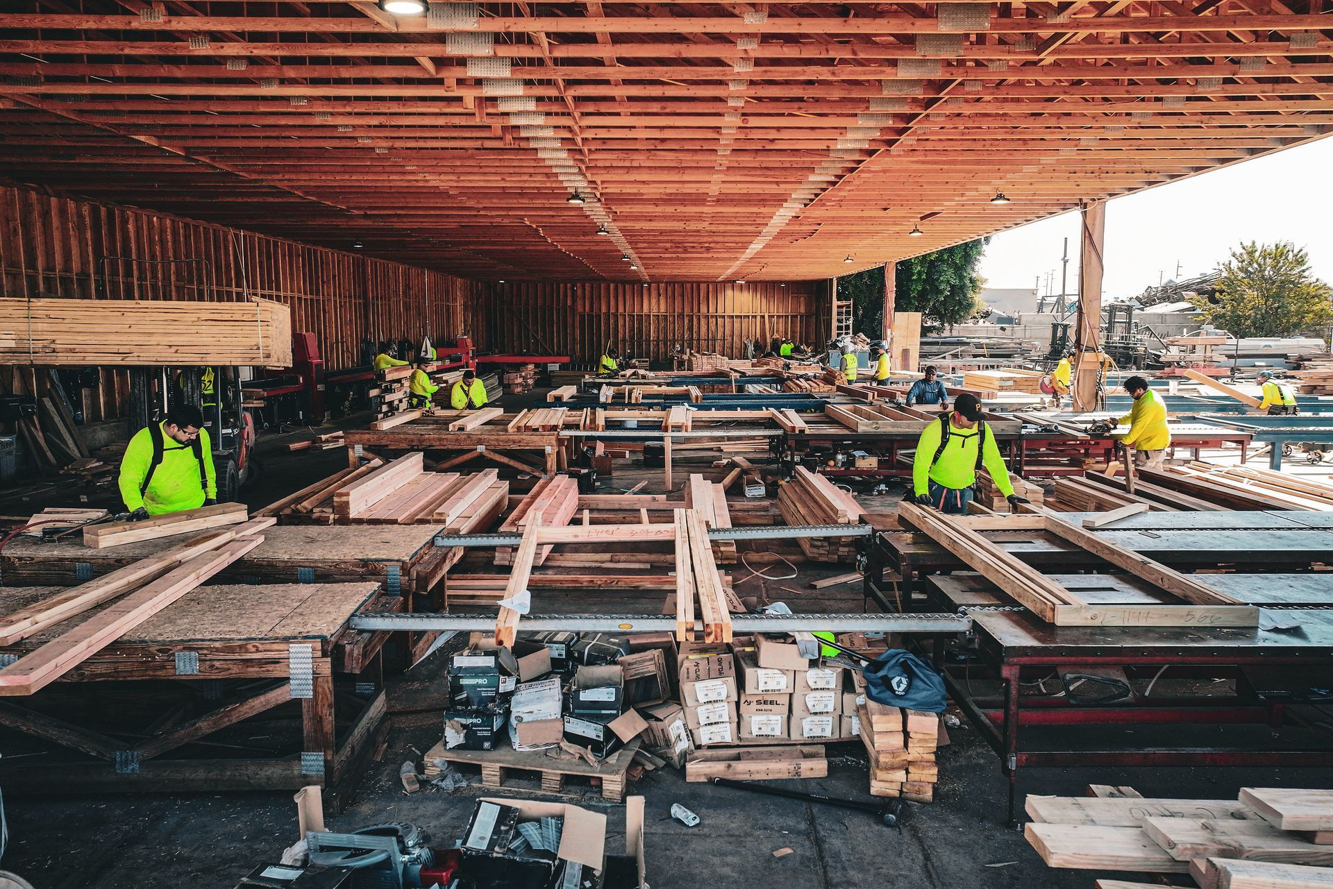 Construction site with wooden framing, stacked materials, and workers in neon safety vests under a roof structure