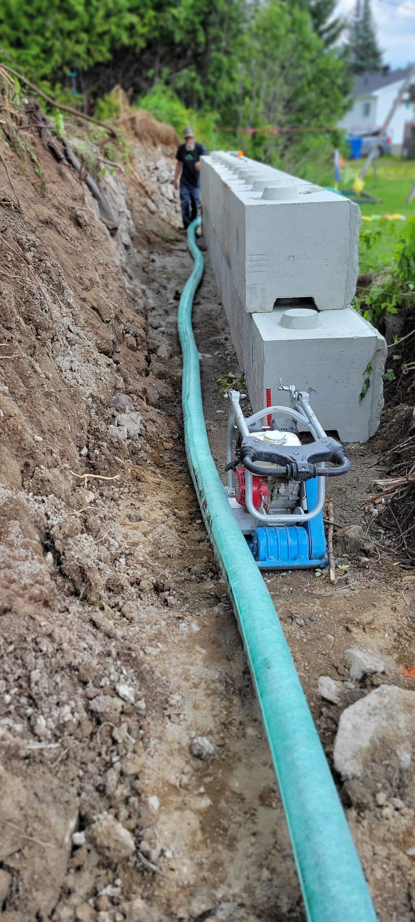 Chantier de construction avec un fossé, un long tuyau vert et des blocs de béton. Une personne travaille.