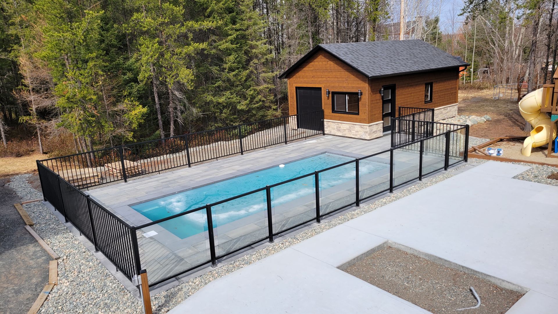 Une piscine rectangulaire entourée d'une clôture noire et d'un petit bâtiment en bois dans un jardin boisé.