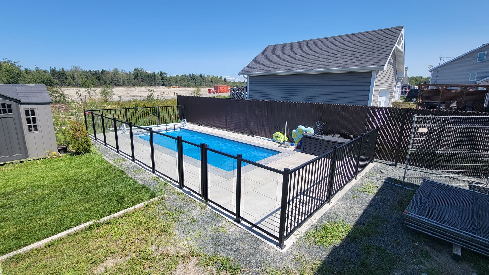 Jardin avec piscine rectangulaire entourée d'une clôture noire. Gazon vert, abri gris et ciel bleu.