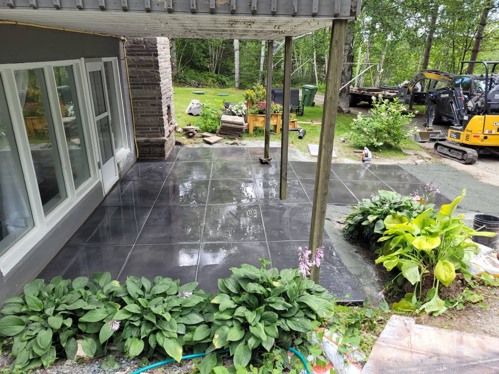 Patio avec carrelage foncé, plantes vertes luxuriantes et arbres en arrière-plan, sous un porche couvert.