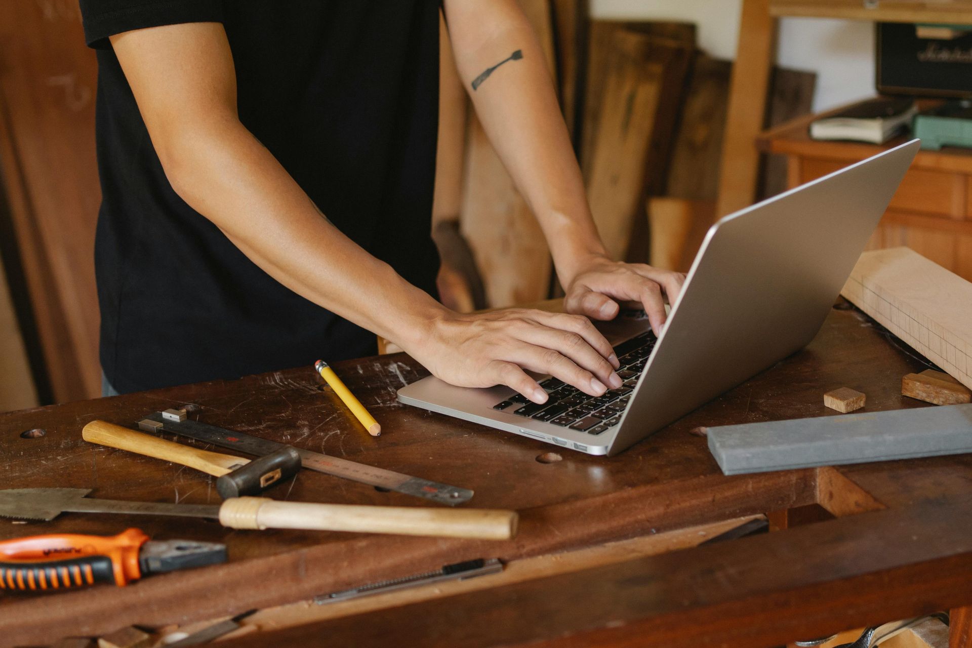 A man is typing on a laptop computer on a wooden table with tools.