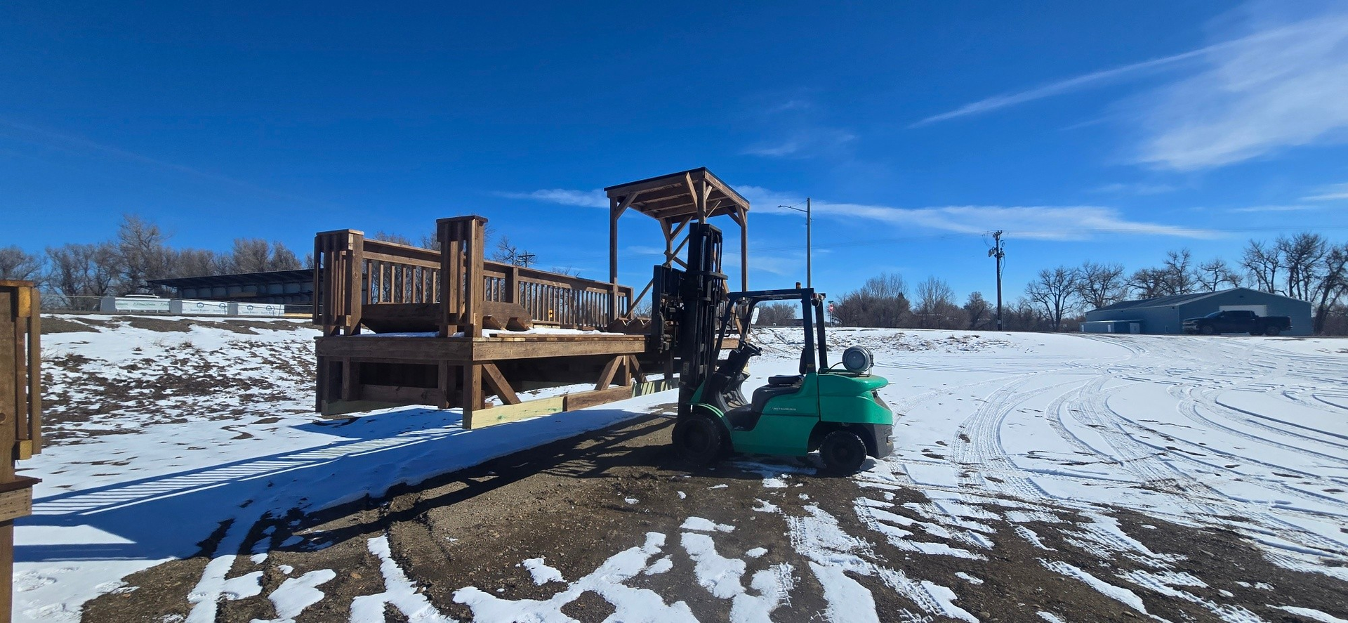 A green forklift lifts a wooden deck structure over a snowy, open field under a clear blue sky.