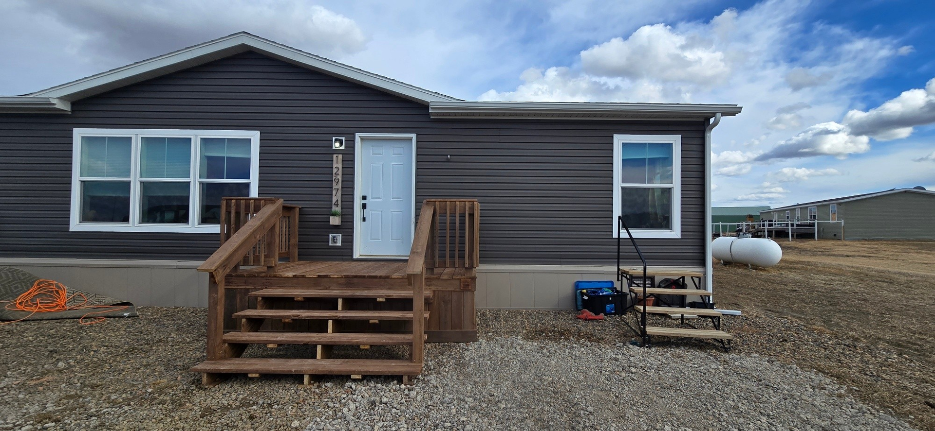 A dark gray manufactured home with a white door, front wood steps, a smaller metal side staircase, and a gravel yard.