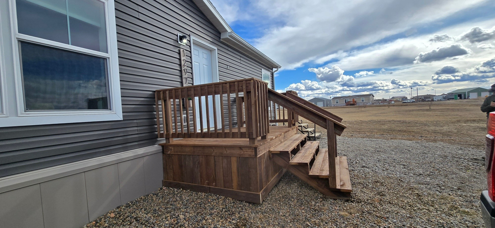 A wooden deck and stairs attached to the side of a grey-sided house, set in an open, gravel-covered area under a cloudy sky.