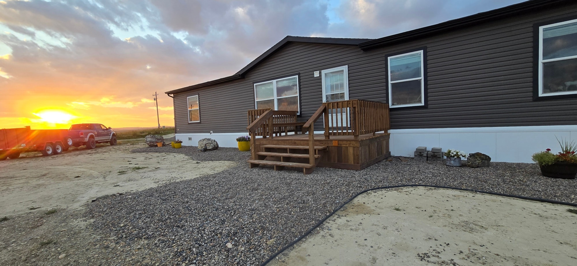 A dark gray manufactured home with a wooden porch at sunset, set against a gravel and dirt yard.